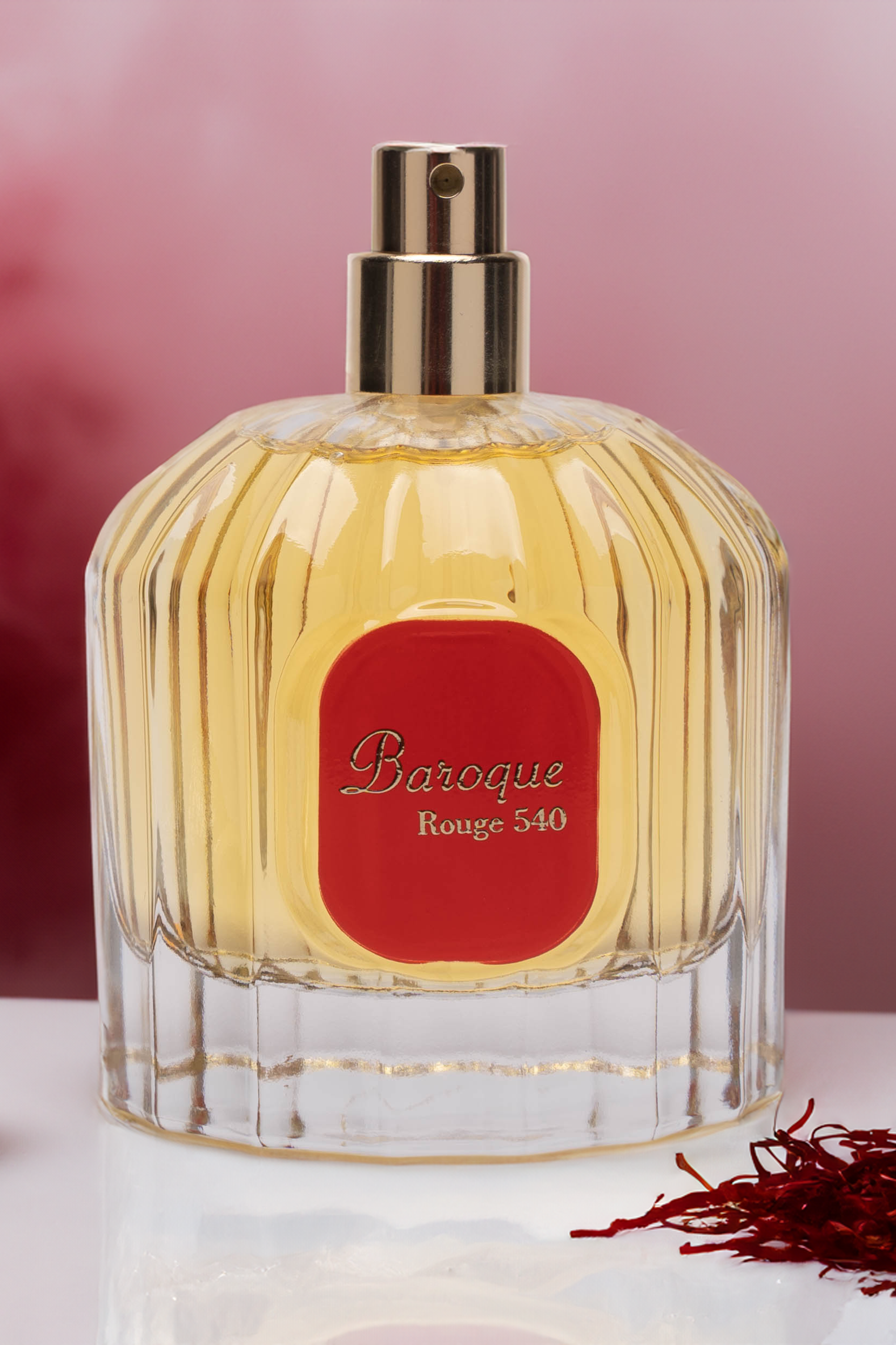 Glass bottle of Maison Alhambra Baroque Rouge 540 perfume, with a vibrant yellow fragrance inside. Decorative saffron is positioned to the right of the bottle, adding a touch of authenticity. The background is enhanced with a deep red mist, emphasizing the luxurious and sophisticated nature of the unisex fragrance